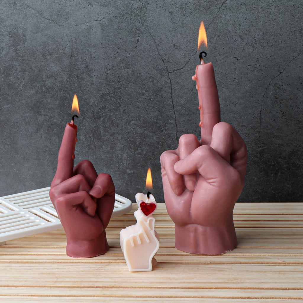 Three novelty hand-shaped candles on a light wood surface against a dark gray textured background. Two larger candles are flesh-toned and show the middle finger with small flames, and one smaller cream candle in front has a red heart detail.
