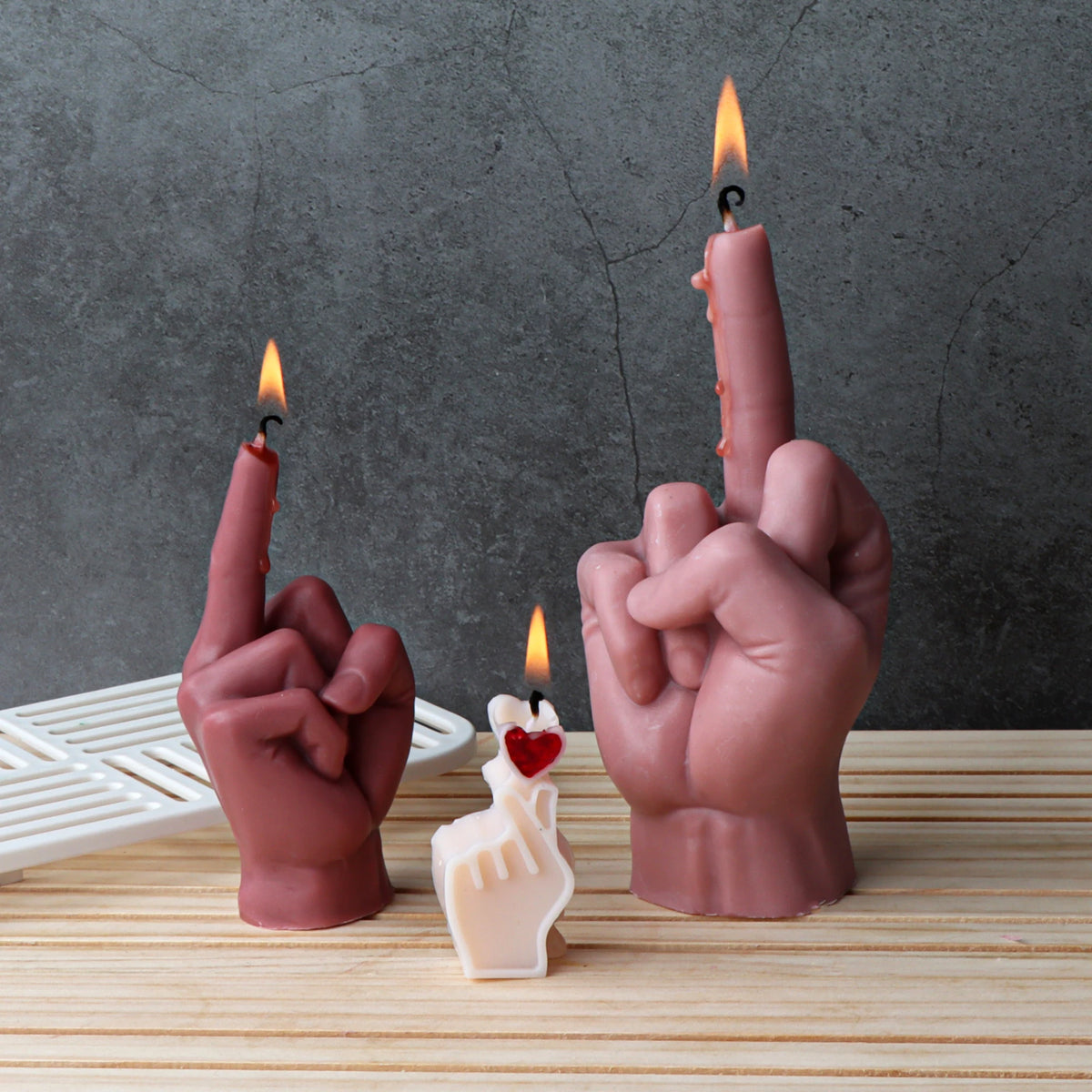 Three novelty hand-shaped candles on a light wood surface against a dark gray textured background. Two larger candles are flesh-toned and show the middle finger with small flames, and one smaller cream candle in front has a red heart detail.
