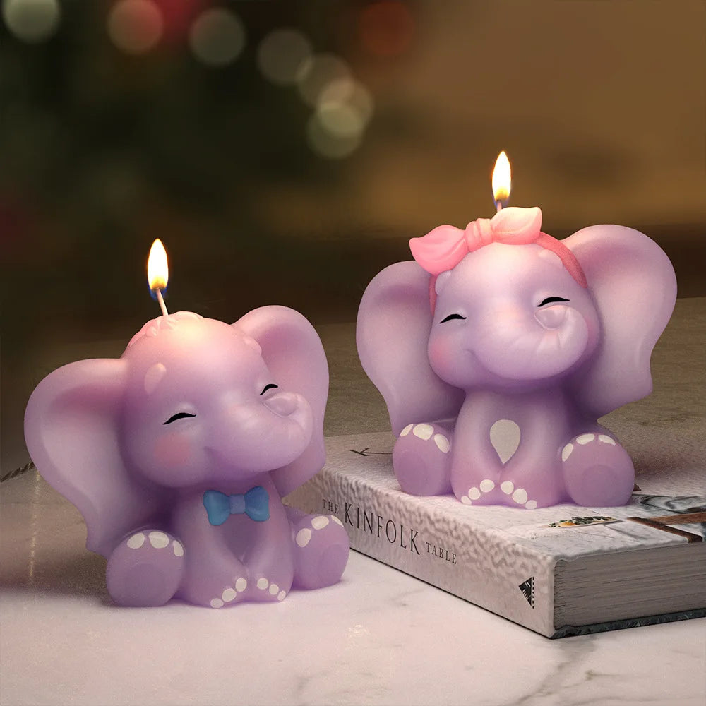 Two lit elephant-shaped candles in pastel purple, one with a blue bow tie and the other with a pink bow, displayed on a tabletop with a blurred festive background.