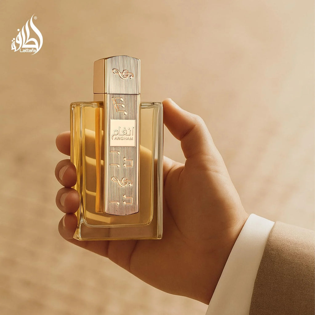 A hand holding a rectangular glass perfume bottle with a metallic silver cap and golden-yellow liquid, set against a warm beige background with a subtle pattern.
