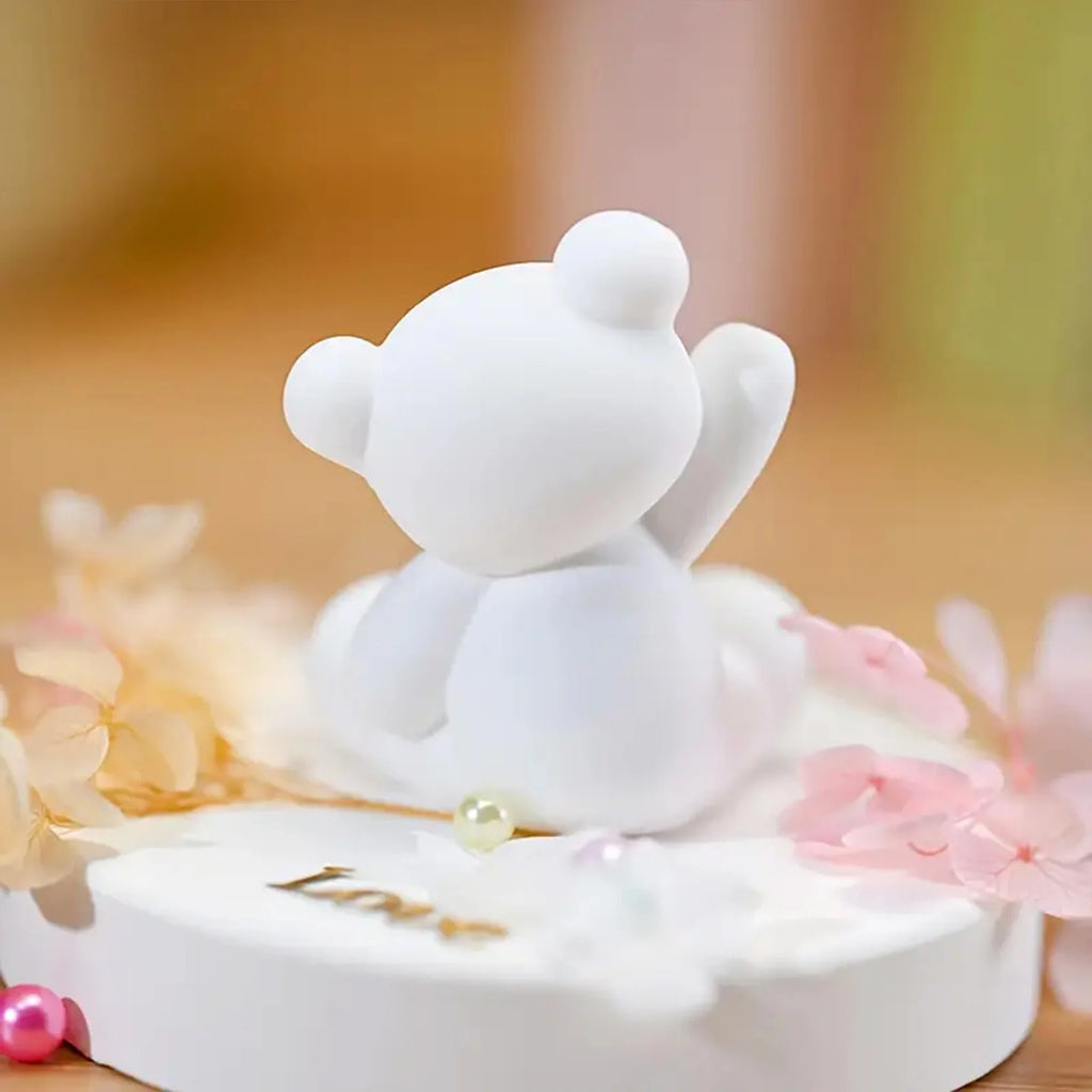 White teddy bear figurine on a white base with pink flowers and lights in the background