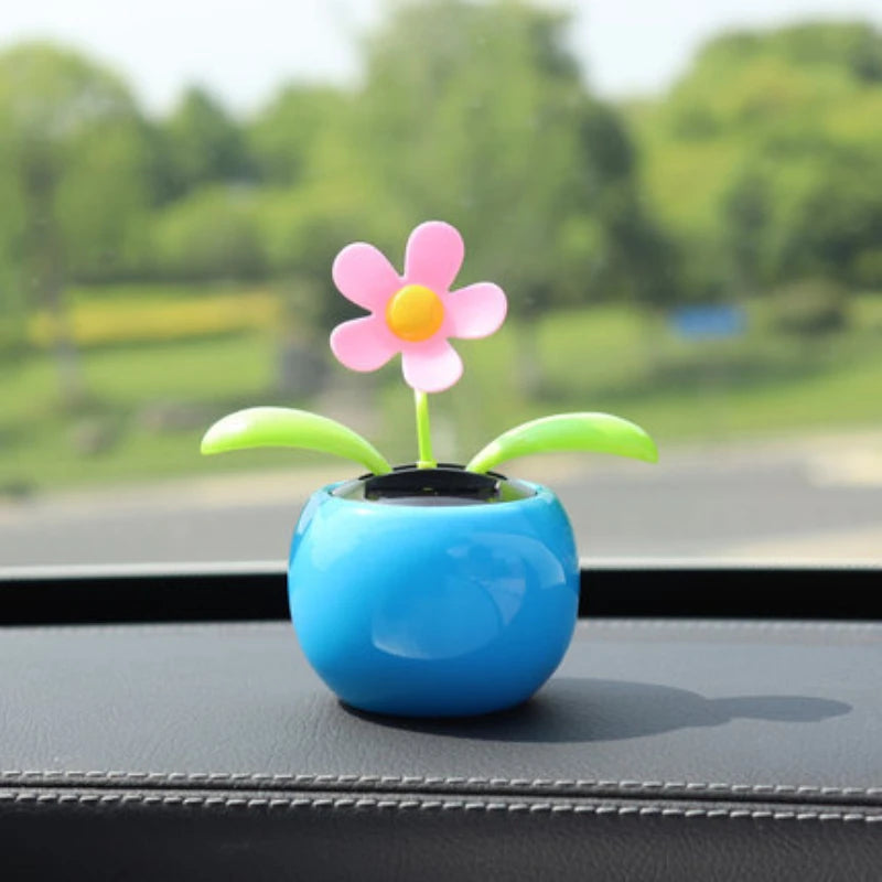 A small decorative car accessory with a blue round pot and a pink flower with green leaves, placed on a car dashboard with a blurred outdoor background visible through the window.