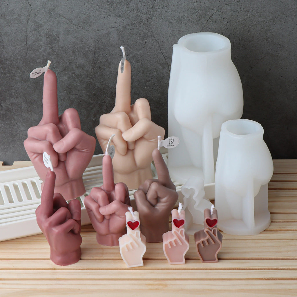 A set of novelty hand-shaped candles and white silicone molds on a light wood surface against a dark gray background. The candles are sculpted in various skin tones and feature a small white heart-shaped charm with a red heart on the index finger.
