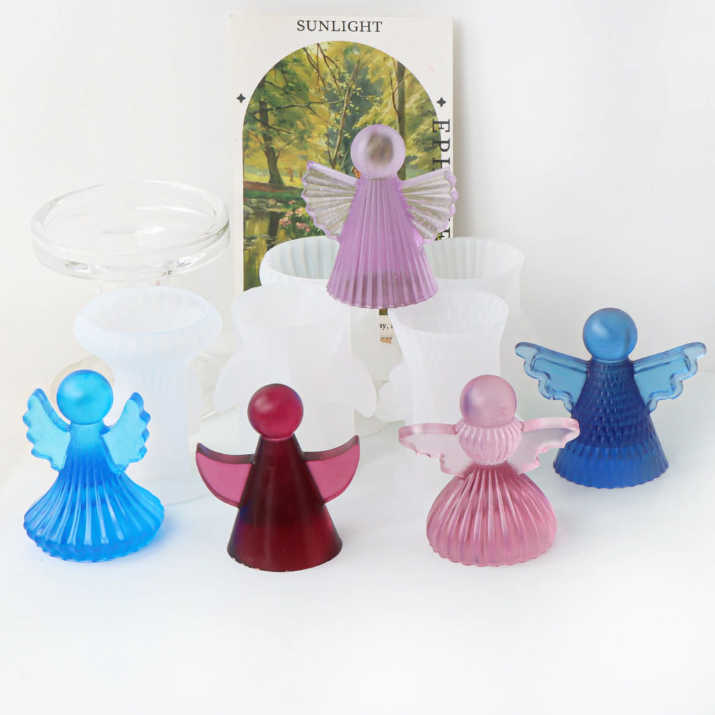 Five small angel-shaped decorative objects in translucent blue, pink, purple, and red are arranged in front of white cups and a card that reads “SUNLIGHT.”.