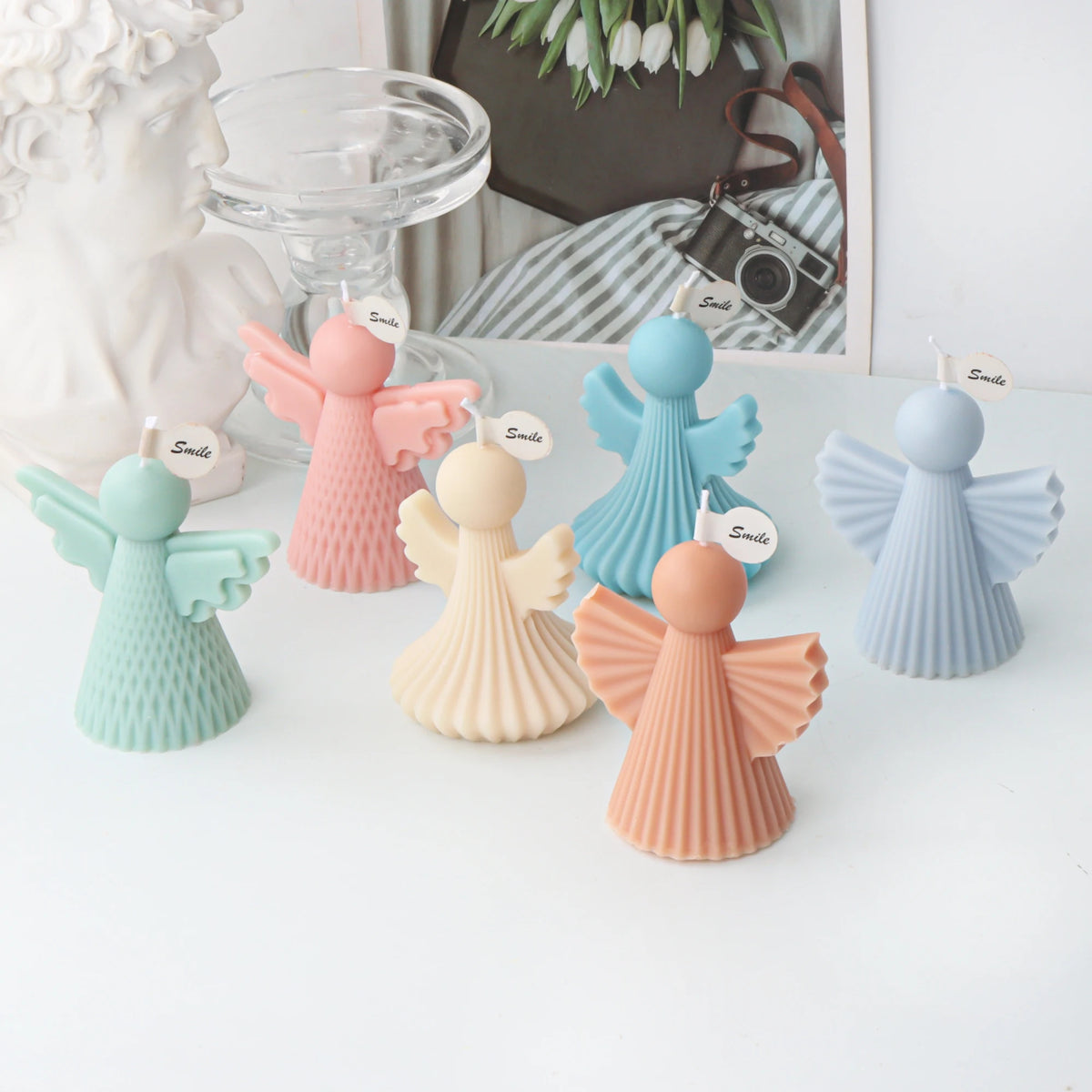 Five angel-shaped candle holders in pastel colors including mint green, pink, light blue, cream, and peach, arranged on a white surface with decorative props in the background.