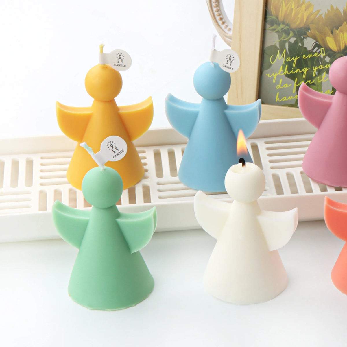 Angel-shaped candle holders in pastel colors including yellow, blue, mint green, white, and pink, arranged on a white surface with one candle lit in the foreground.