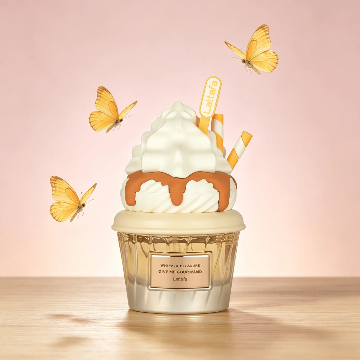 A Lattafa Perfume cupcake-shaped perfume bottle on a wooden surface against a soft pink background, decorated with white whipped cream, caramel drizzle, striped straws, and yellow butterflies. The bottle label reads 'Whipped Vanilla Give Me Courrègo'.