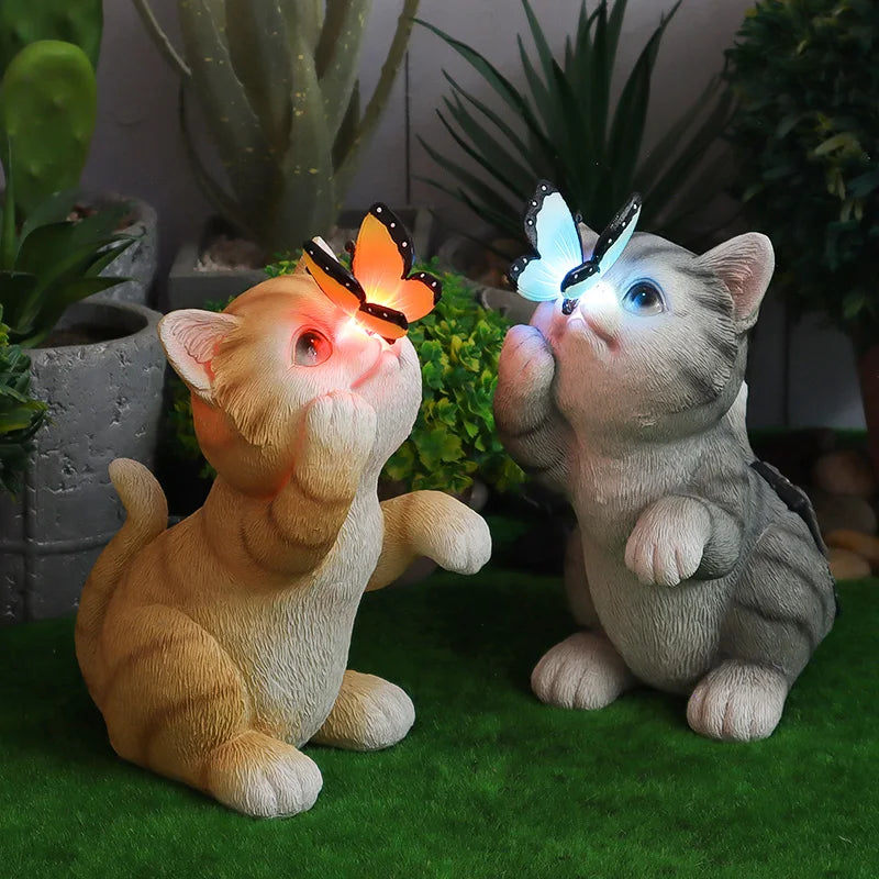 Two small cat garden statues posed on artificial green grass, each with a glowing butterfly above its face. One cat is cream/tan and the other is gray, with the left cat's butterfly emitting orange light and the right cat's butterfly emitting blue light.