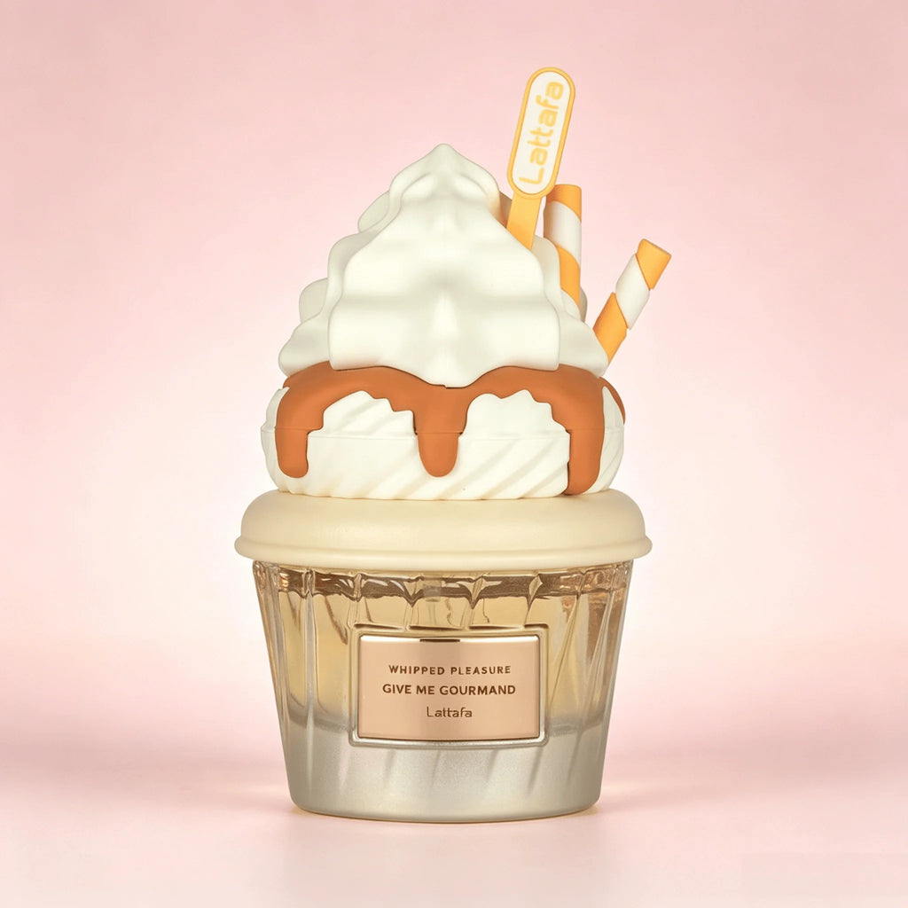 A Lattafa Perfume novelty candle shaped like a whipped cream dessert cup, with white whipped cream, brown caramel drizzle, a clear faceted cup base, and two orange-and-white striped straws, on a soft pink background.