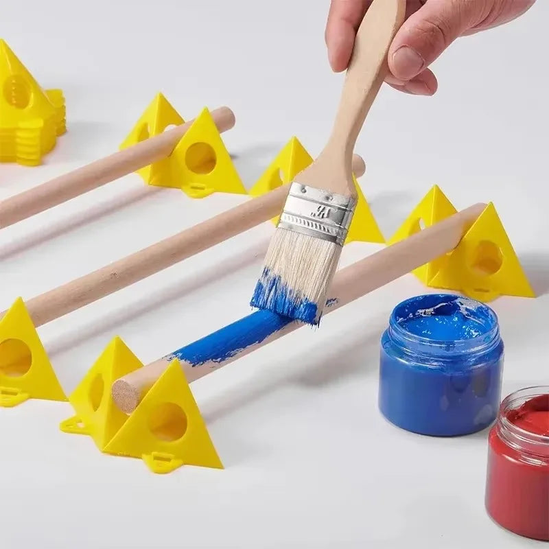 A hand holds a paintbrush applying blue paint to a wooden dowel, with yellow triangular pegs, additional wooden rods, blue and red paint jars, and more paintbrushes arranged on a white surface.