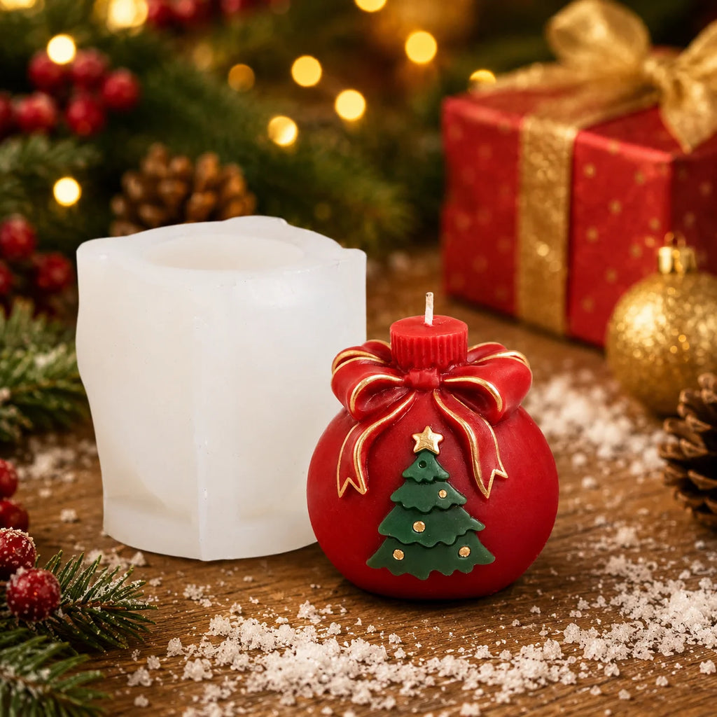 A Christmas candle mold set on a wooden surface, featuring a red ornament-shaped candle with a green Christmas tree and gold bow, next to a white wax candle, with festive holiday decorations and wrapped gifts in the background.