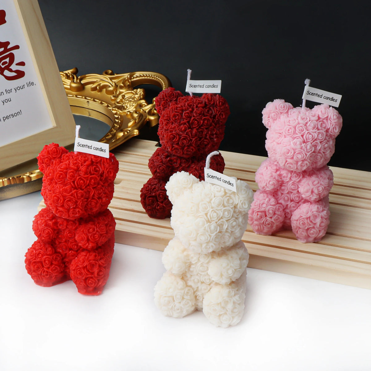 Four rose-textured teddy bear candles in red, dark red, ivory and pink beside a gold-colored mirror