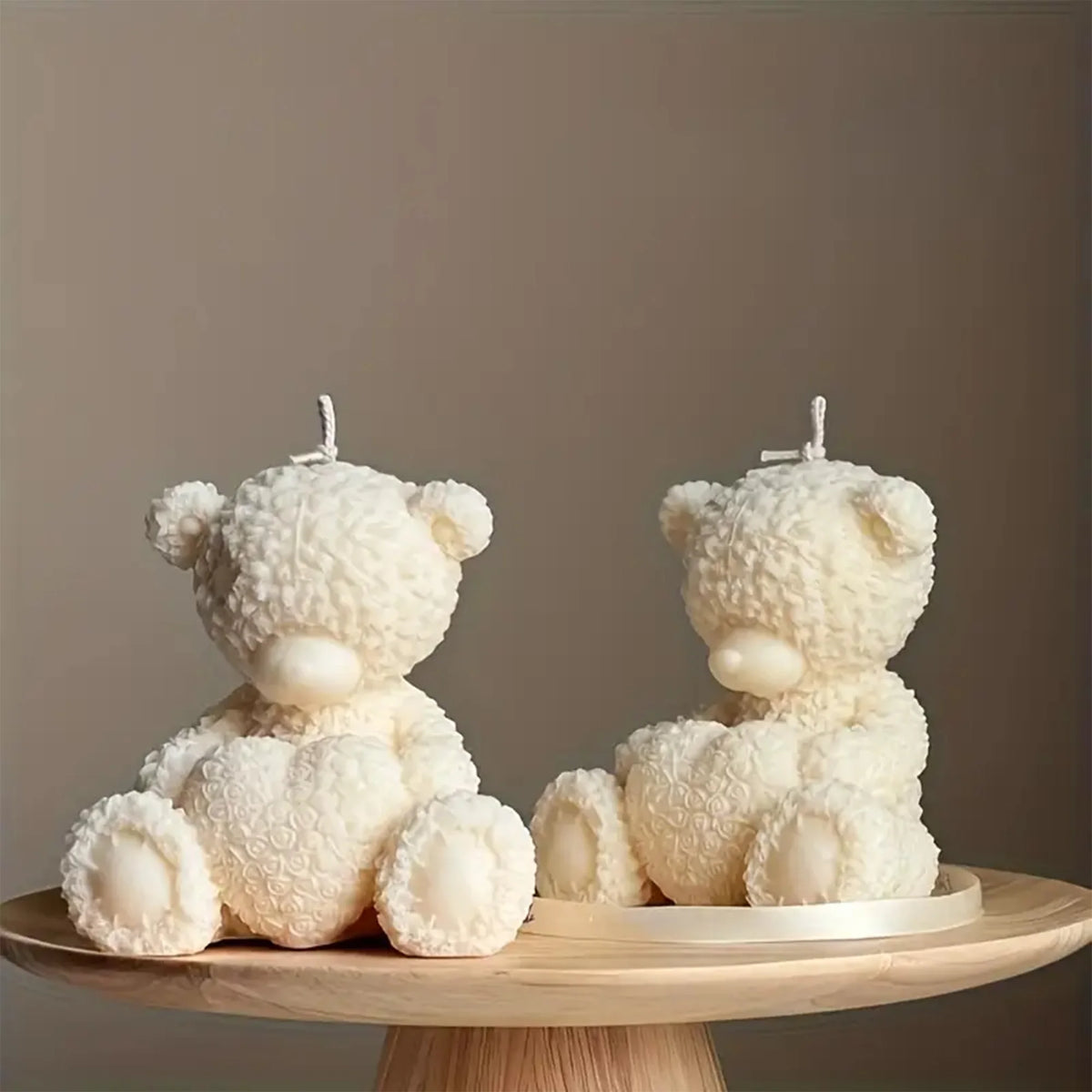 Two ivory seated teddy bear example candles holding textured hearts on a round wooden stand