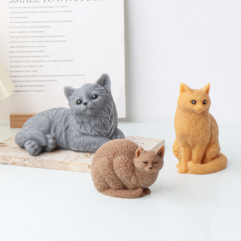Three realistic candle mold cat figurines in gray, orange, and brown, displayed on a white surface with a book in the background.