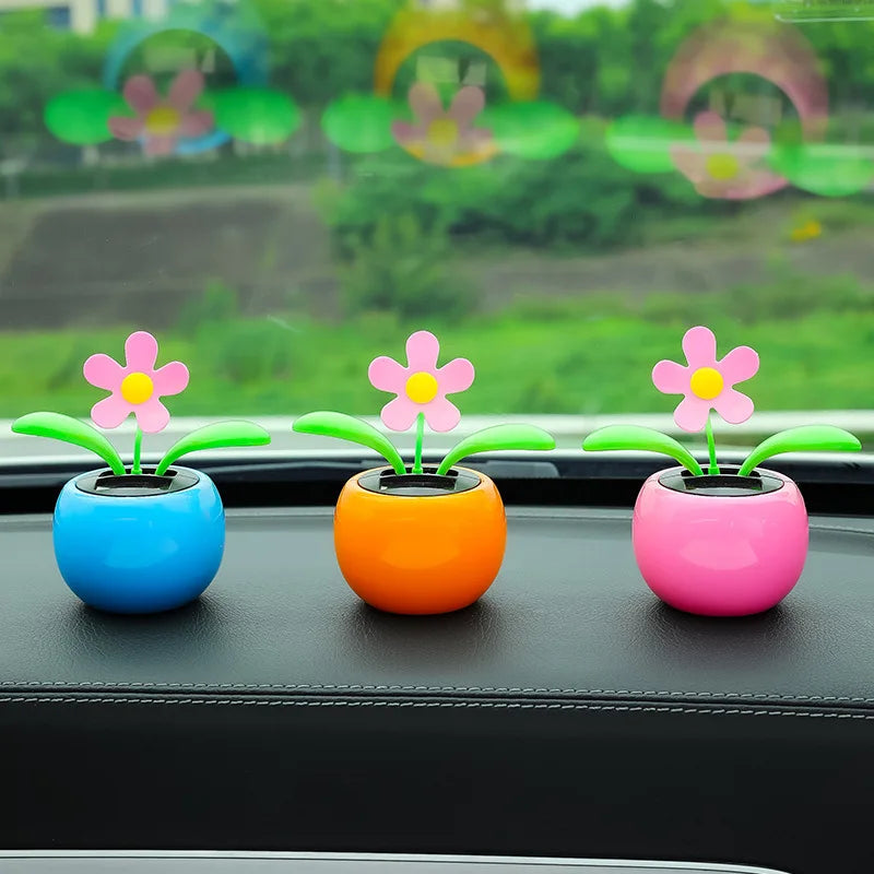 Three small flower-shaped car air fresheners in blue, orange, and pink pots, placed on a black car dashboard with a blurred green outdoor background visible through the windshield.