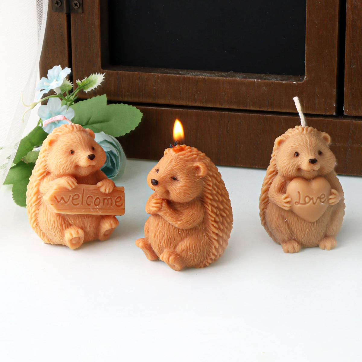 Three tan hedgehog example candles, including Welcome sign and Love heart designs, with the middle candle lit