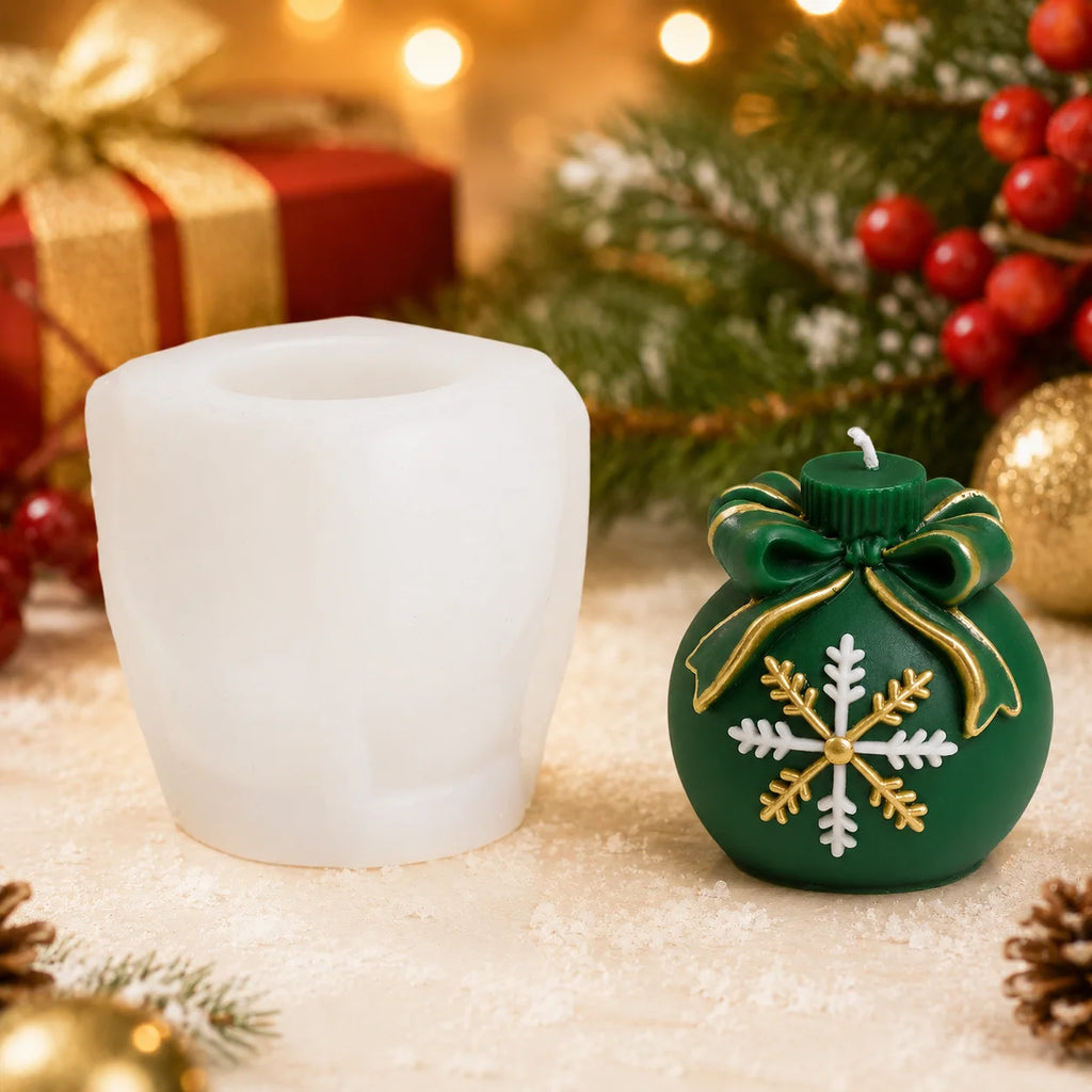 A holiday-themed product display featuring a christmas candle mold and a green ornament-shaped candle decorated with a gold and white snowflake and green bow, set against a festive Christmas background with pine branches, red berries, and warm bokeh lights.