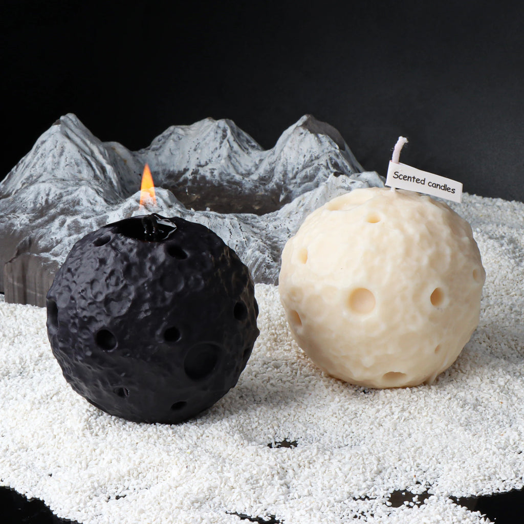 Black and ivory crater-textured moon candles, one lit, against a miniature lunar landscape