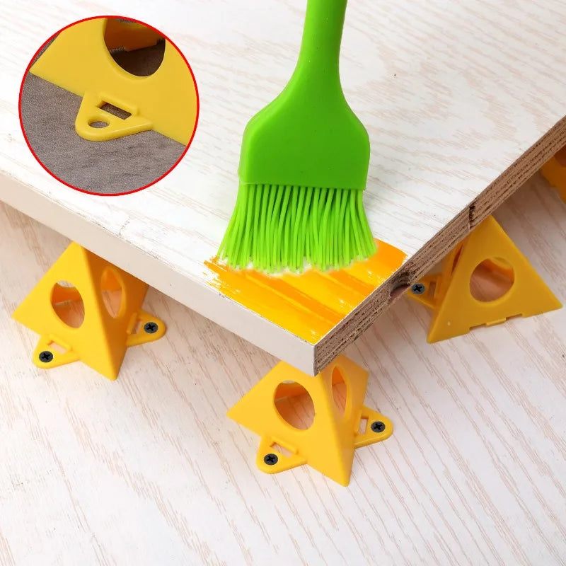 Yellow triangular leveling feet supporting a white wooden board on a light wood floor, with a green paint brush applying yellow paint along the edge of the board. A circular inset shows a close-up of one leveling foot.