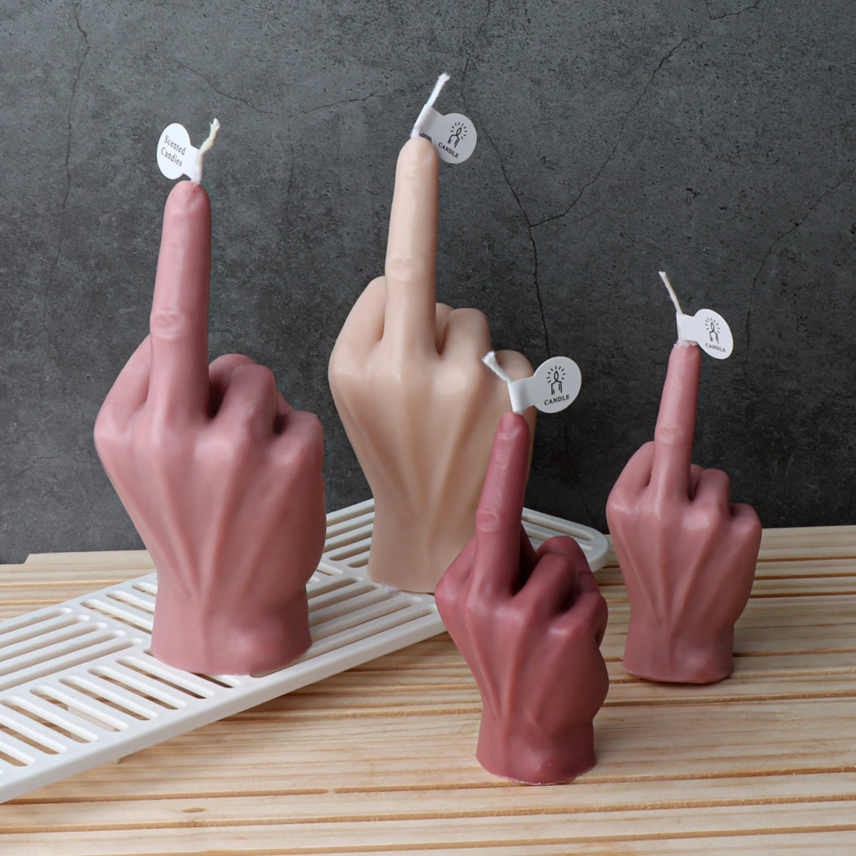 Three novelty hand-shaped candles shaped like middle fingers in dusty rose, beige, and mauve tones, arranged on a white slatted tray over a light wood surface against a dark textured wall.