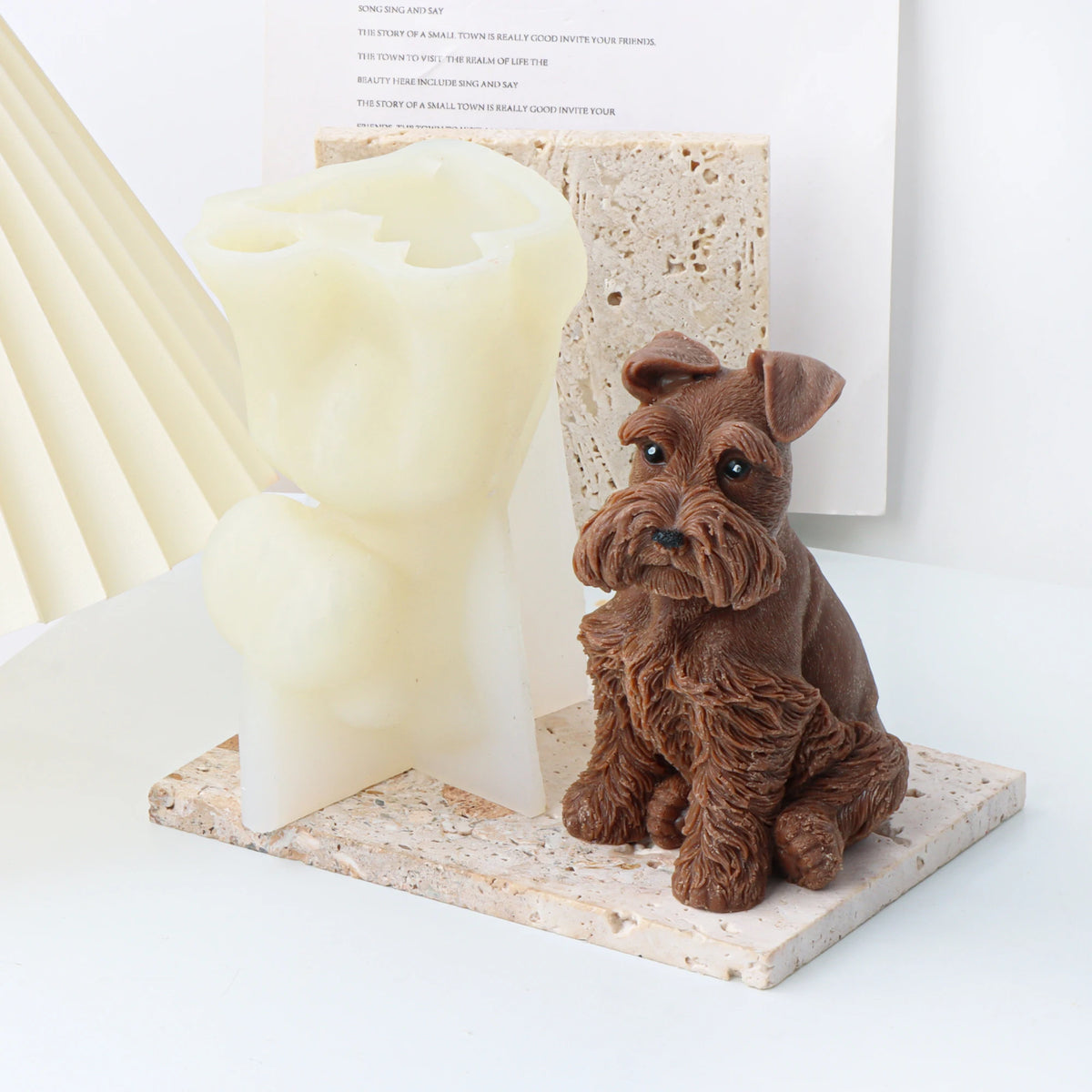 A decorative scene with a small brown dog candle mold figurine sitting on a stone-look base next to a translucent cream-colored sculptural object, with a stone tile and a book in the background.