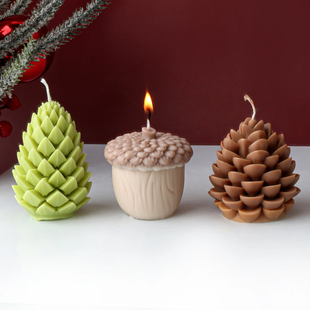 Three decorative candles shaped like a green pine cone, acorn, and brown pine cone on a white surface with a red background.