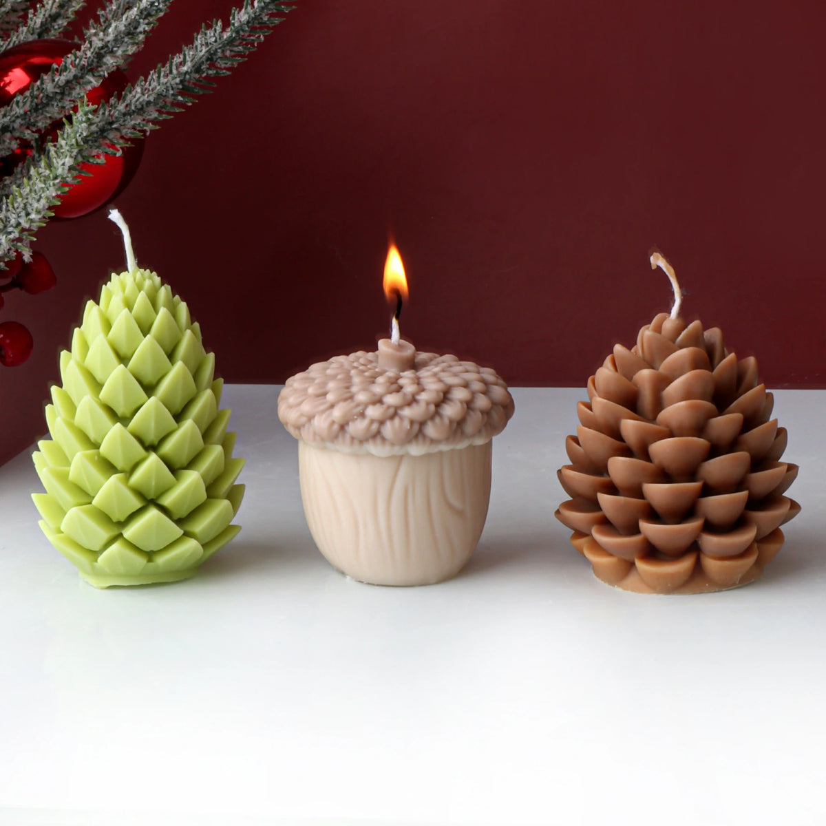 Three decorative candles shaped like a green pine cone, acorn, and brown pine cone on a white surface with a red background.