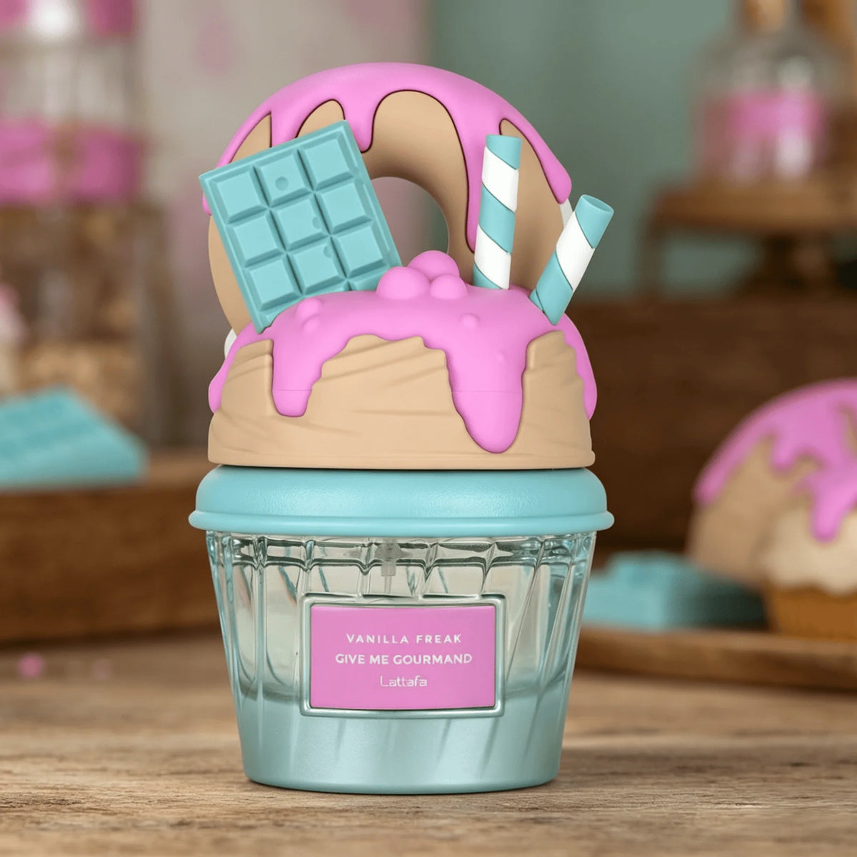 A cupcake-shaped Lattafa perfume atomizer labeled 'Vanilla Freak Gys Me Gourmand Lattafa' in light blue, topped with pink frosting, a donut, a chocolate bar, and striped straws, set on a wooden surface with a blurred bakery background.