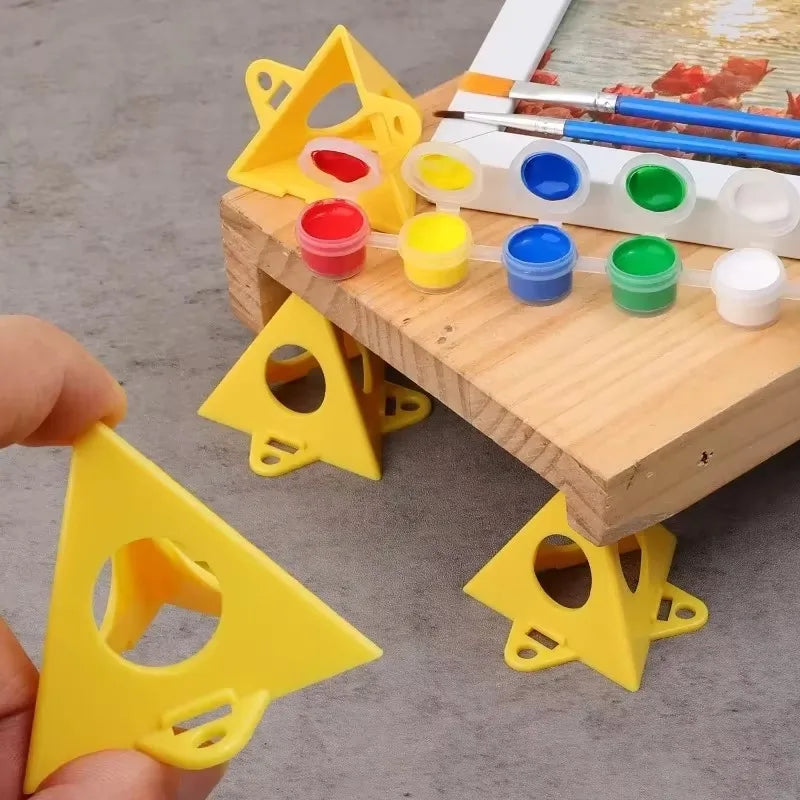 Yellow plastic triangular leveling blocks with circular cutouts, shown being held by a hand and placed under a wooden easel with paint pots and brushes on a gray floor.