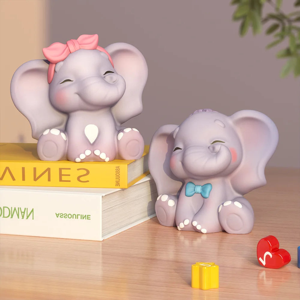 Two small elephant figurines on a wooden surface, one pink and one lavender with a blue bow tie, styled with books and small heart-shaped blocks.
