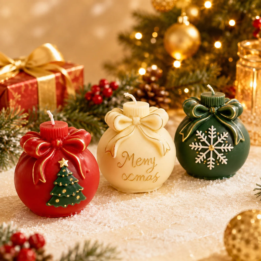 Three Christmas ornament-shaped candles are displayed on a snowy surface with festive decorations in the background. The left candle is red with a gold bow and a small green Christmas tree design.