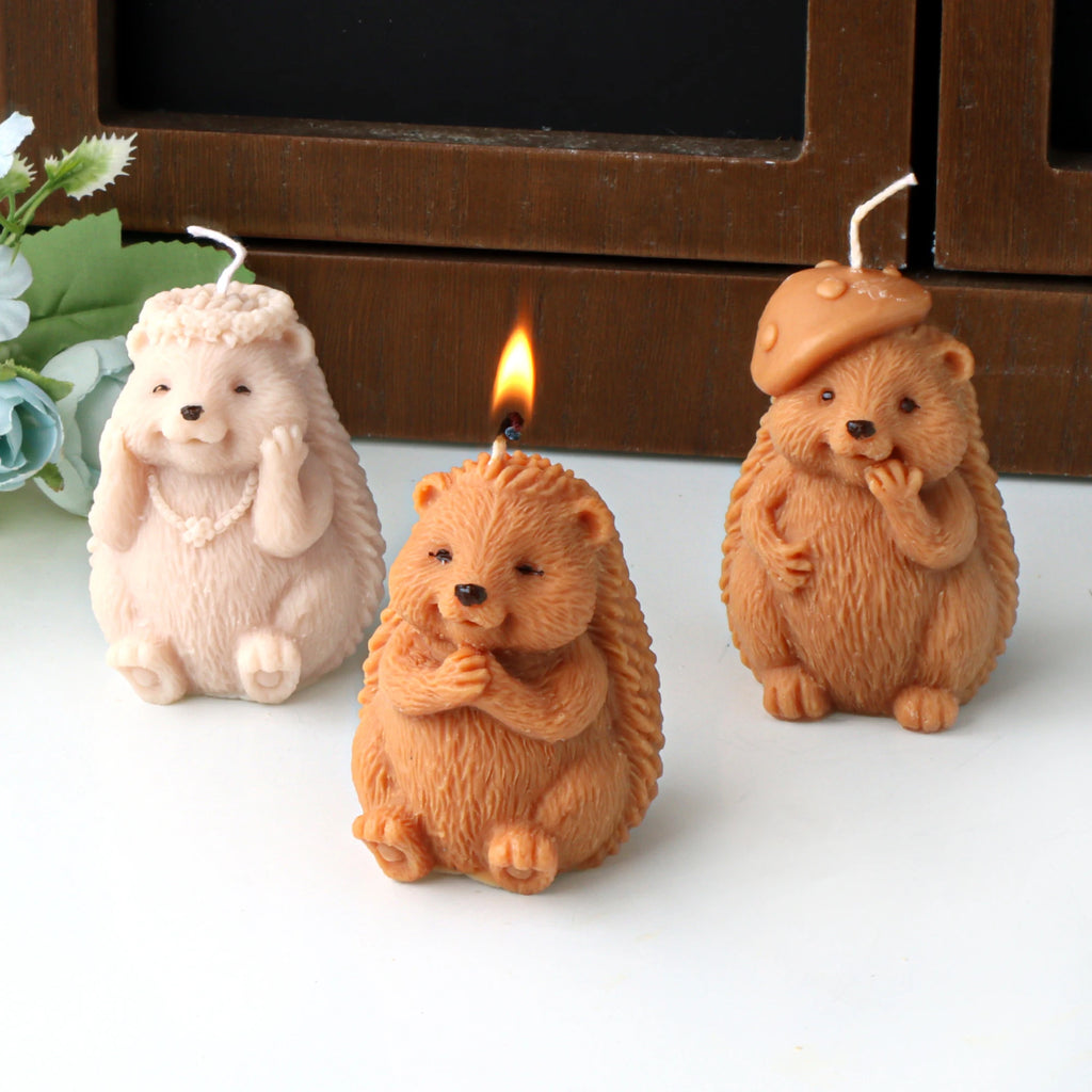Three hedgehog example candles with flower-crown, hands-together and mushroom-cap designs, the center one lit