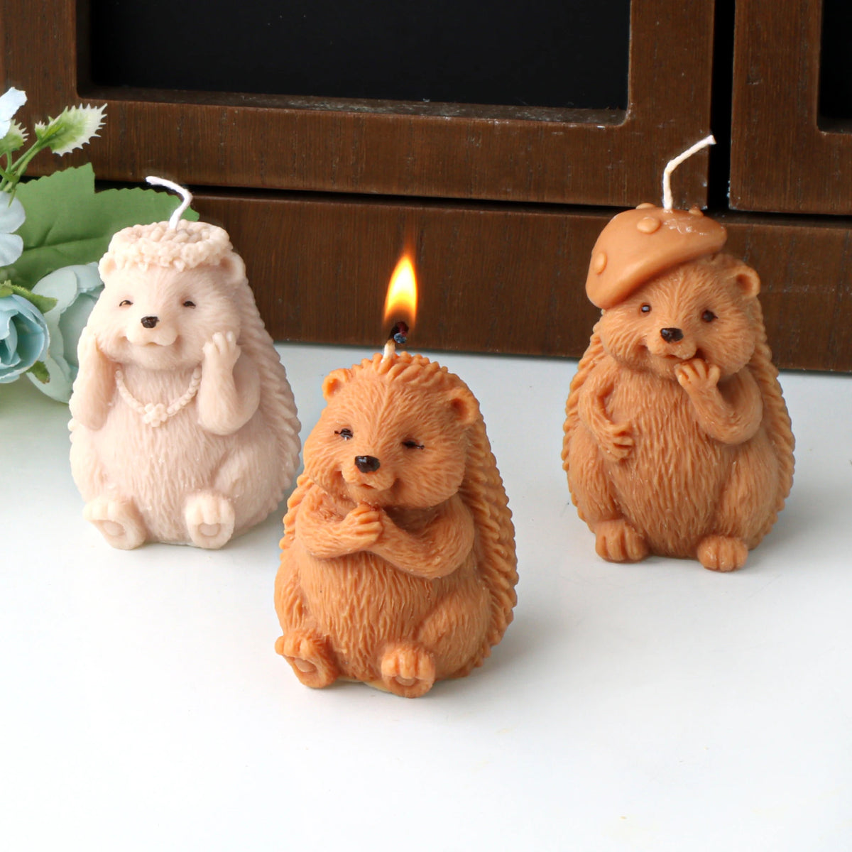 Three hedgehog example candles with flower-crown, hands-together and mushroom-cap designs, the center one lit