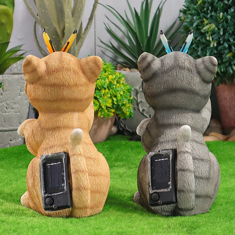 Two cat-shaped garden statues shown from the back on artificial grass, each with a small solar panel on the back and colored pencils sticking out of the ears. One cat is tan and the other is gray, with potted plants in the background.