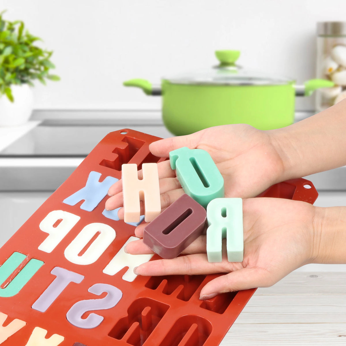 Hands holding four colored letter casting examples above a red alphabet silicone mold