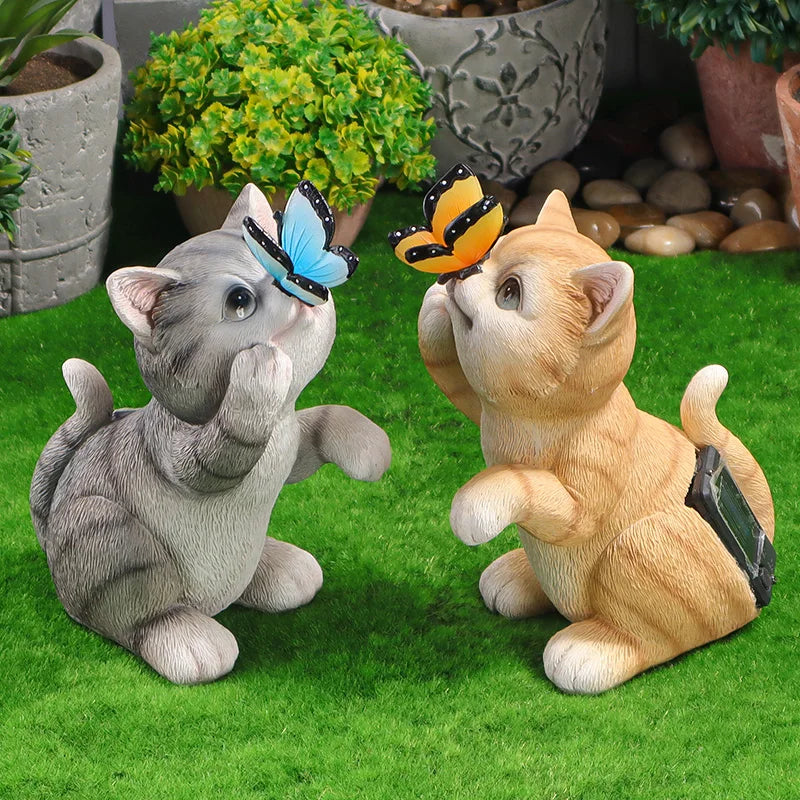 Two realistic cat garden statues posed facing each other on artificial grass, with one gray cat holding a blue butterfly and one tan cat holding an orange-and-black butterfly. Decorative potted plants appear in the background.
