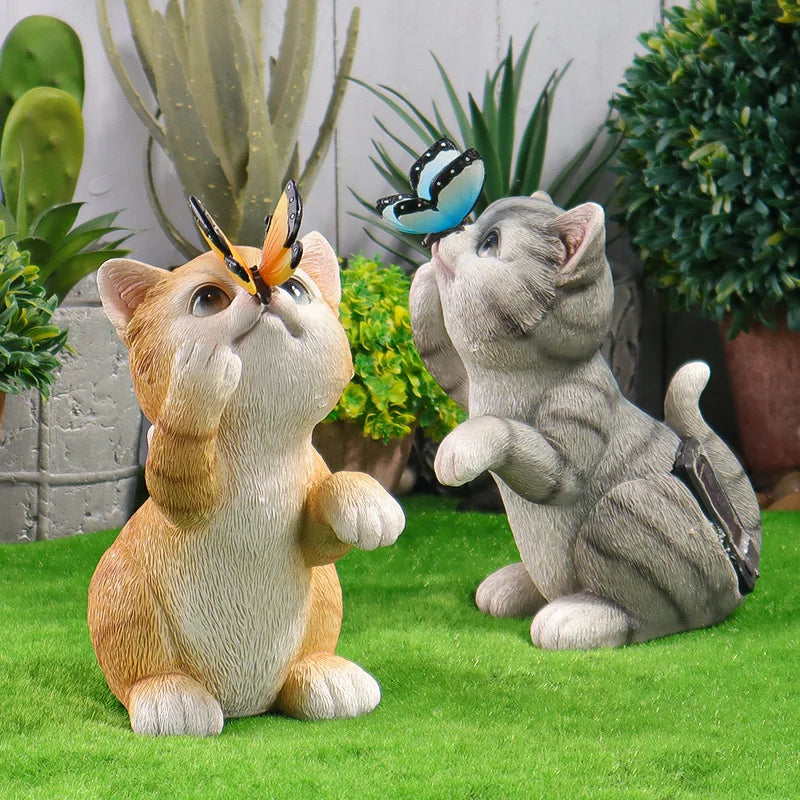 Two funny cat garden statues posed on artificial grass with potted plants in the background. One cat is orange and white with a butterfly on its head, and the other is gray with a butterfly on its back.